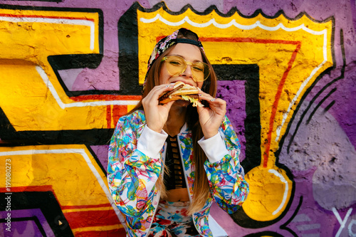 Beautiful cheerful girl, eating a tasty burger, takes pleasure of food. Dressed in bright sportive suit and cap, in sunglasses. Standing outdoors, near the wall with graffiti.