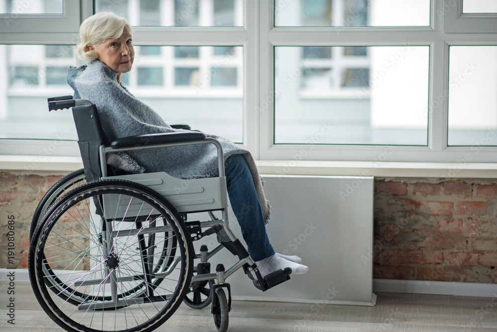 Upset mature woman looking at camera with sad look. She is sitting in wheelchair in room wrapped in a blanket. Copy space in right side Upset mature woman looking at camera with sad look. She is sitting in wheelchair in room wrapped in a blanket. Copy space in right side