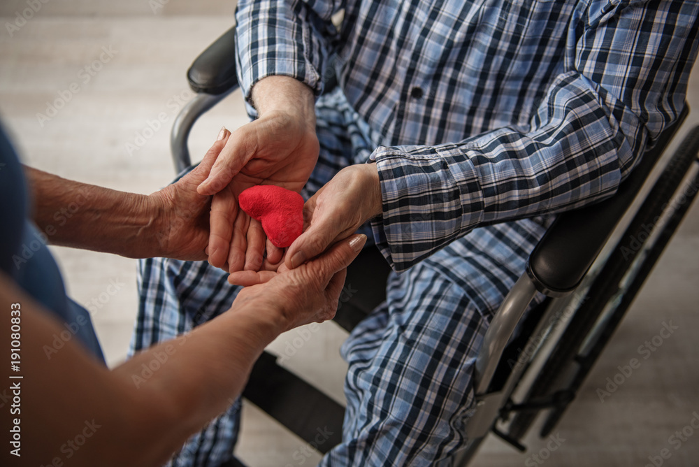 Top view close up of male and female hands holding red small toy heart. Man is sitting in invalid chair. Woman is standing near him. Concept of love