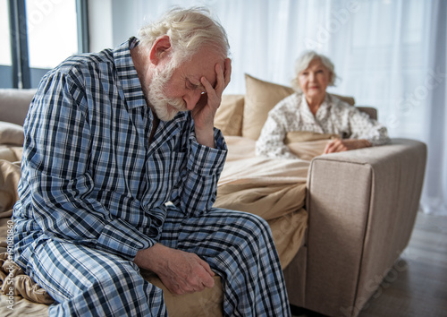 Feeling bad. Moody old husband sitting on bed with sad look and propping head with his hand. His wife sitting in bed and looking at him