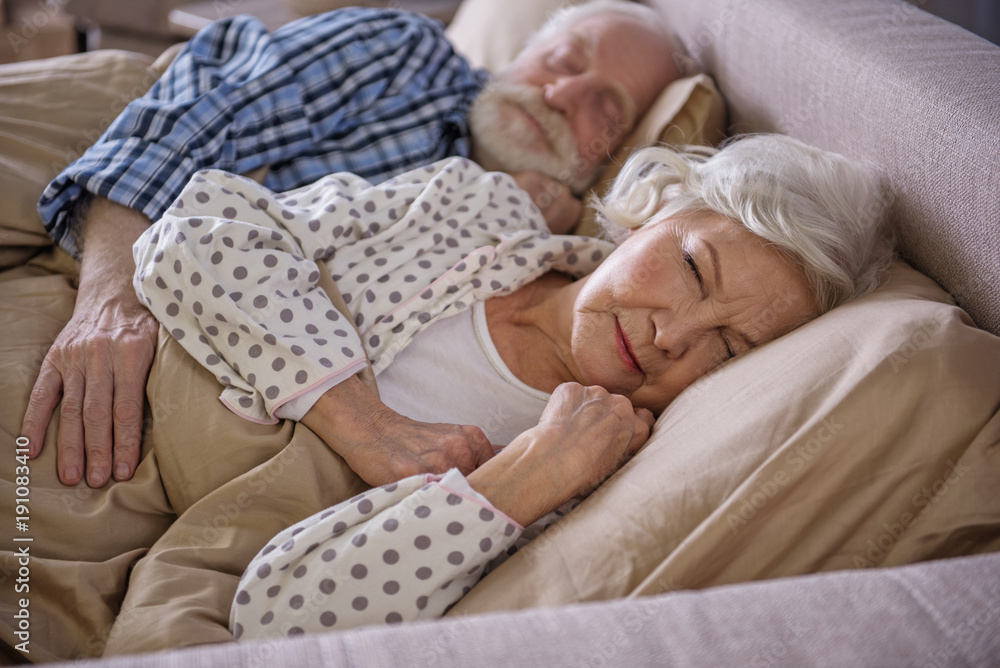 Tranquil married couple lying in bed and sleeping. Old husband is embracing his wife while they are having rest at home Tranquil married couple lying in bed and sleeping. Old husband is embracing his wife while they are having rest at home