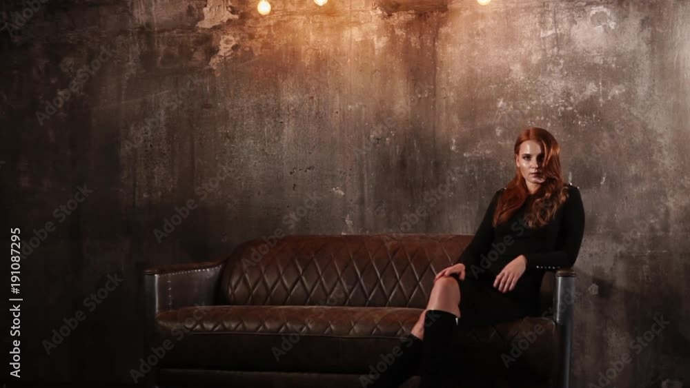 Amazing model with a red hair is queitly sitting on brown leather couch. She brushes her hairs back and smiles. Amazing model with a red hair is queitly sitting on brown leather couch. She brushes her hairs back and smiles.