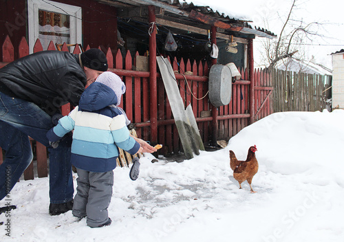 A man with a little boy, feeding a chicken in a village yard, in winter