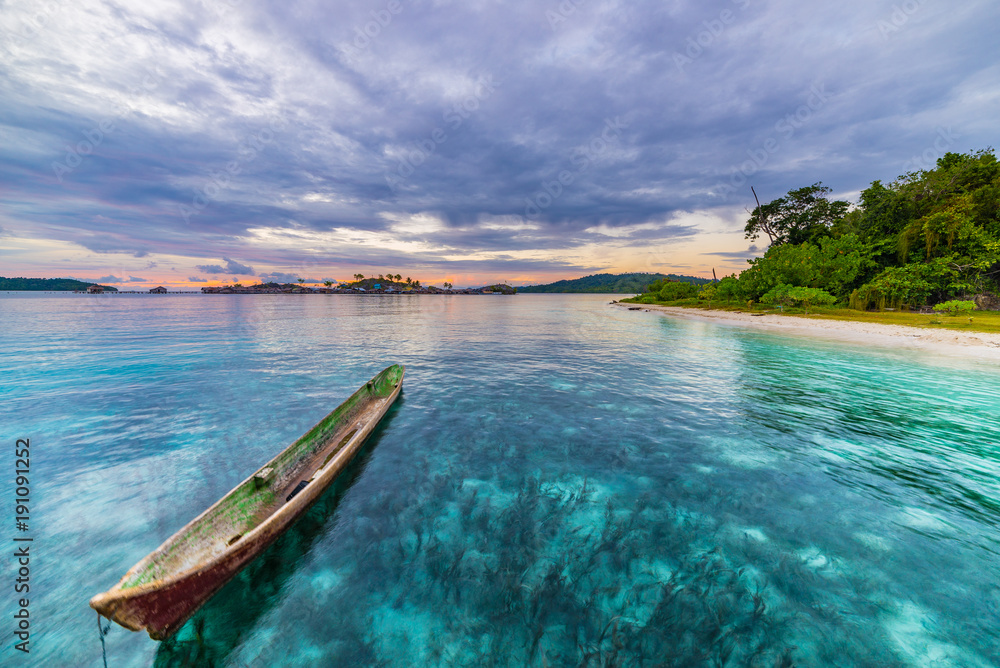 Tropical beach, caribbean sea, canoe floating on transparent turquoise water, remote Togean Islands (Togian Islands), Sulawesi, Indonesia. Dramatic sky at sunset.