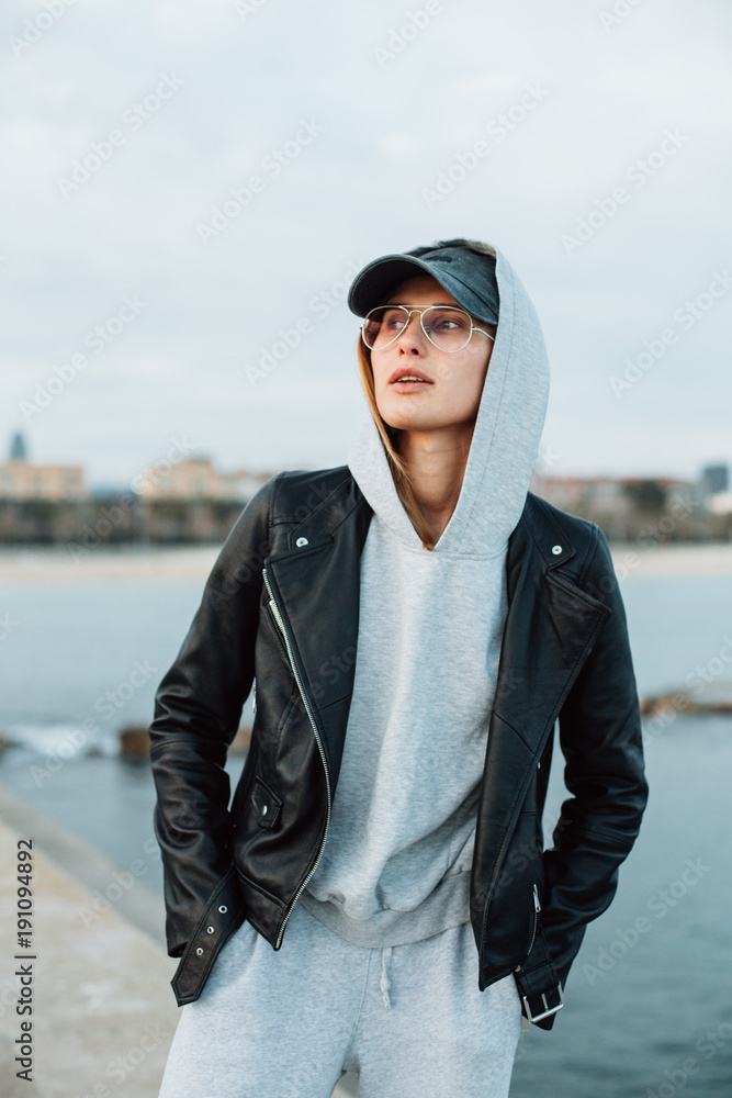 Hip and stylish trendy young hipster millennial woman in street style fashion look and outfit wears baseball cap and leather jacket stands on beach seafront at sunset confident and independent Stock P...