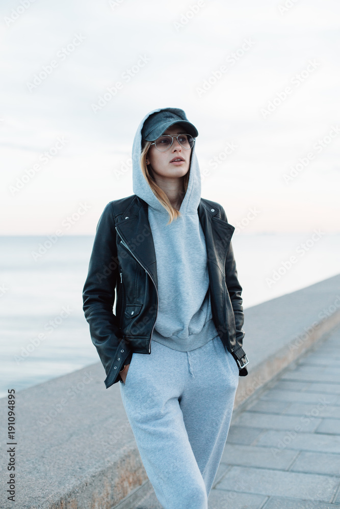 Hip and stylish trendy young hipster millennial woman in street style fashion look and outfit, wears baseball cap and leather jacket, stands on beach seafront at sunset confident and independent Hip and stylish trendy young hipster millennial woman in street style fashion look and outfit, wears baseball cap and leather jacket, stands on beach seafront at sunset confident and independent