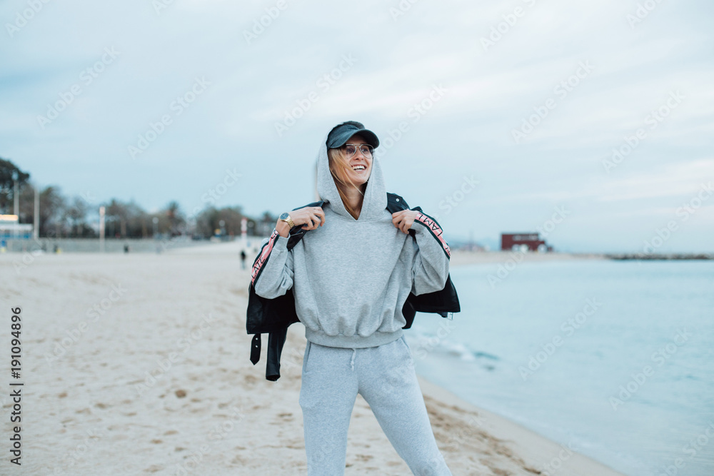Happy and natural beauty young pretty and attractive woman or teenager wears grey hoodie and sweatpants during workout at beach seaside laughs naturally to camera, during fashion photoshoot influencer Happy and natural beauty young pretty and attractive woman or teenager wears grey hoodie and sweatpants during workout at beach seaside laughs naturally to camera, during fashion photoshoot influencer