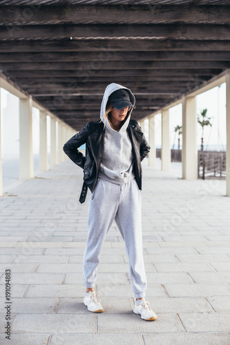 Cool and hip young female in grey fashionable tracksuit, sweatpants and hoodie poses for camera during fashion photoshoot, urban street style for social media or fashion outfit blog photo