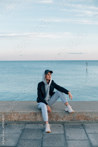 Trendy and cool young female in aviator glasses, baseball cap, sweatpants and hoodie sits on concrete pier next to sea or ocean,poses for camera with model face expression, hip and beautiful empowered