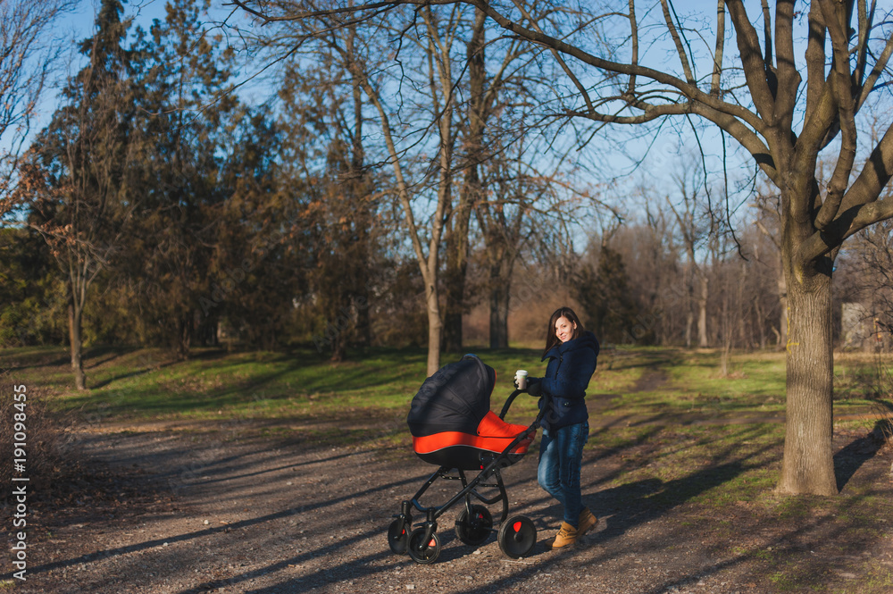 Young woman in blue coat walking in autumn park with little child baby in orange baby carriage, tea or coffe on fall nature, green trees. Mother and kid. Parenthood, love, parents, children concept. Young woman in blue coat walking in autumn park with little child baby in orange baby carriage, tea or coffe on fall nature, green trees. Mother and kid. Parenthood, love, parents, children concept.