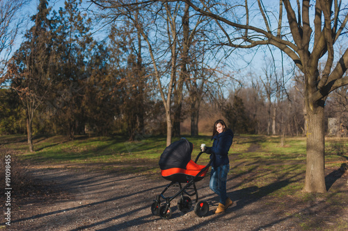 Young woman in blue coat walking in autumn park with little child baby in orange baby carriage, tea or coffe on fall nature, green trees. Mother and kid. Parenthood, love, parents, children concept.