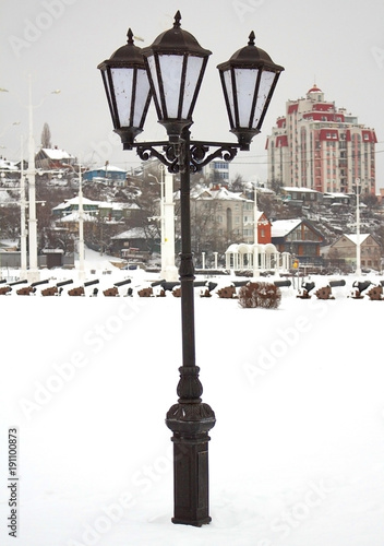 Iron, forged lantern, black color stands on the city street, on a winter day in the snow