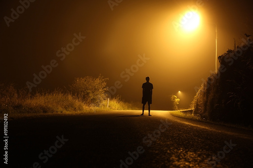 The silhouette of a man in shorts, standing in the middle of the road on a misty night. The glare of the street light against the fog sets a creepy mood. Concept of thieves, rapist and killers roaming