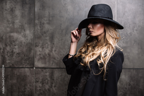 Beautiful young mysterious blond girl in black hat and black jacket on gray background. Eyes are covered with a hat. Manicure - long red nails, nail polish. Fashion, beauty. © boykovi1991