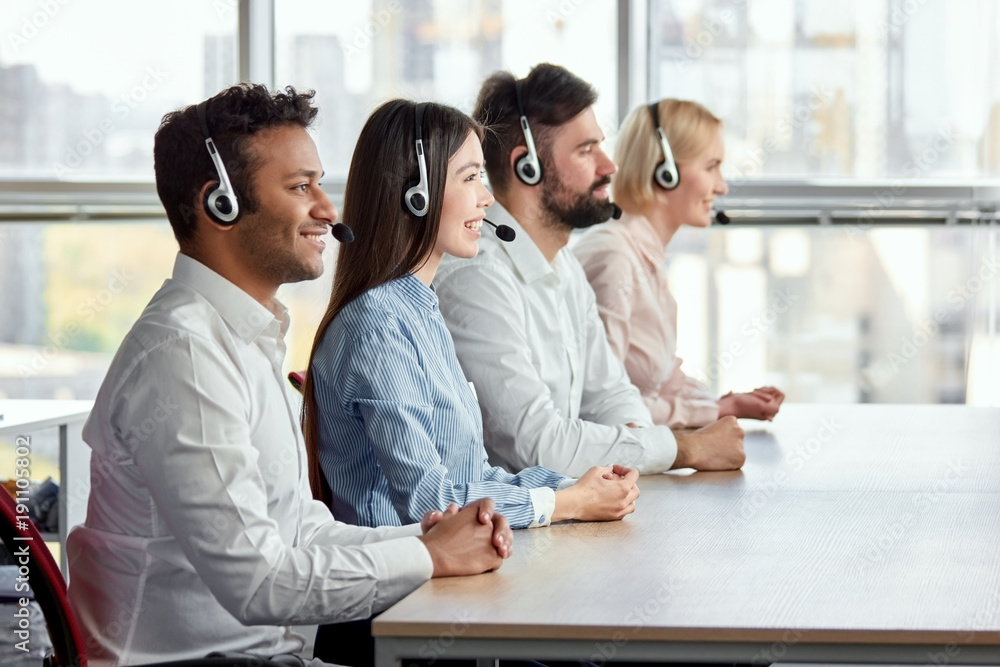 Side view happy multicultural call center operators sitting at desk. Call center workers with folded hands in bright office. Side view happy multicultural call center operators sitting at desk. Call center workers with folded hands in bright office.