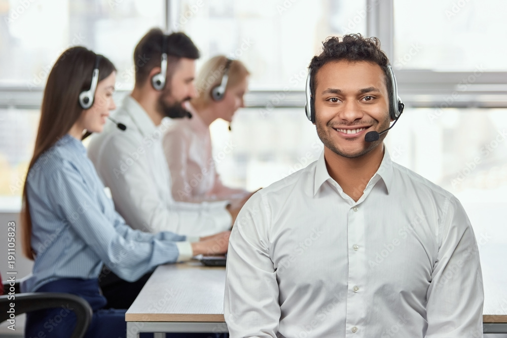 Young friendly male customer service operator. Portrait of a smiling businessman with headsets in a call centre. Bright windows background. Young friendly male customer service operator. Portrait of a smiling businessman with headsets in a call centre. Bright windows background.