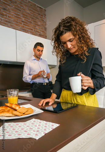 Curly woman looking electronic tablet while young man having fast coffee in home before go to work