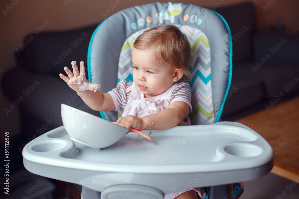 Portrait of cute adorable Caucasian child kid girl sitting in high chair eating cereal with spoon. Everyday lifestyle. Candid real authentic moment Portrait of cute adorable Caucasian child kid girl sitting in high chair eating cereal with spoon. Everyday lifestyle. Candid real authentic moment