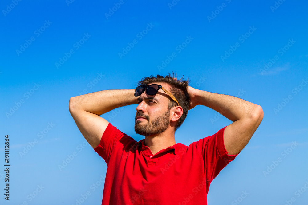 Dynamic, ambitious, successful, expressive bearded young in red t-shirt sunglasses in summer blue sky ocean background. Self-confidence lifestyle concept Dynamic, ambitious, successful, expressive bearded young in red t-shirt sunglasses in summer blue sky ocean background. Self-confidence lifestyle concept