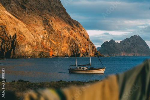 View on a white sailing boat anchored in natural harbour in a secret bay close to Desertas islands during sunset with orange sunny mountains in the background and wet clothes hanged in foreground  photo