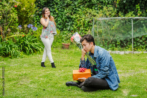 Close up of sad man wearing a jean jacket and black pants sitting in the ground holding gift with a blurred woman behind using her cellphone. Valentine's Day concept or friend zone concept
