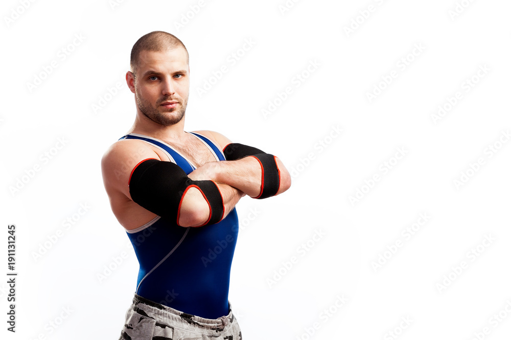 Young athletic man wrestler in a blue vest, sports pants and elbow pads stands with arms crossed on a white isolated background, side view