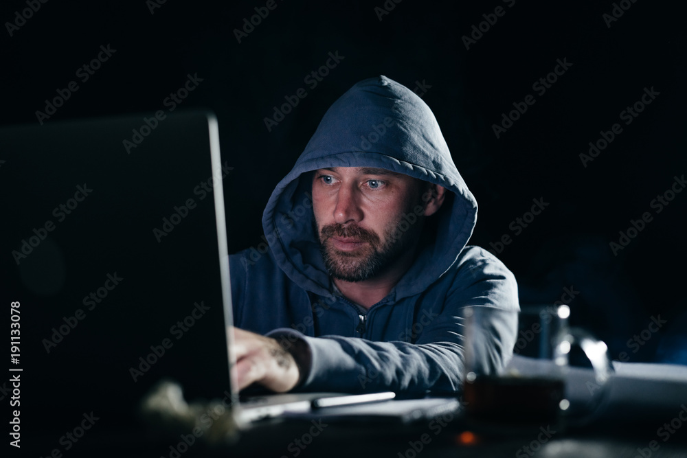 male hacker in a sweatshirt with a hood sitting and looking at the laptop male hacker in a sweatshirt with a hood sitting and looking at the laptop