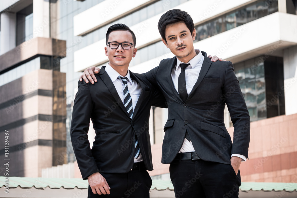 Portrait of two young Asian businessmen in black suit standing with office building background Portrait of two young Asian businessmen in black suit standing with office building background