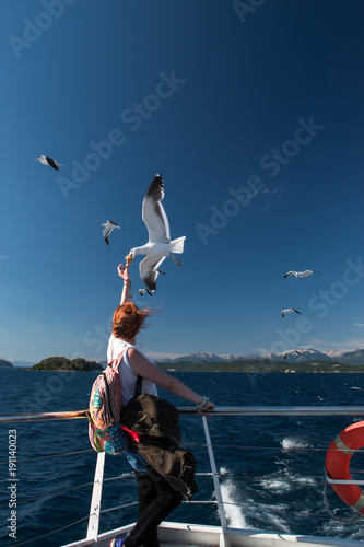 Chica de pelo colorado alimentando a una gaviota en un bote, con lago de fondo en dia despejado