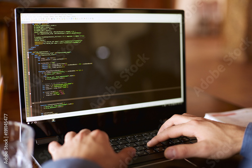 Back view over the shoulder shot of developer programmer with laptop. Program code and script data on the screen. Young freelancer in glasses working on project in cafe.