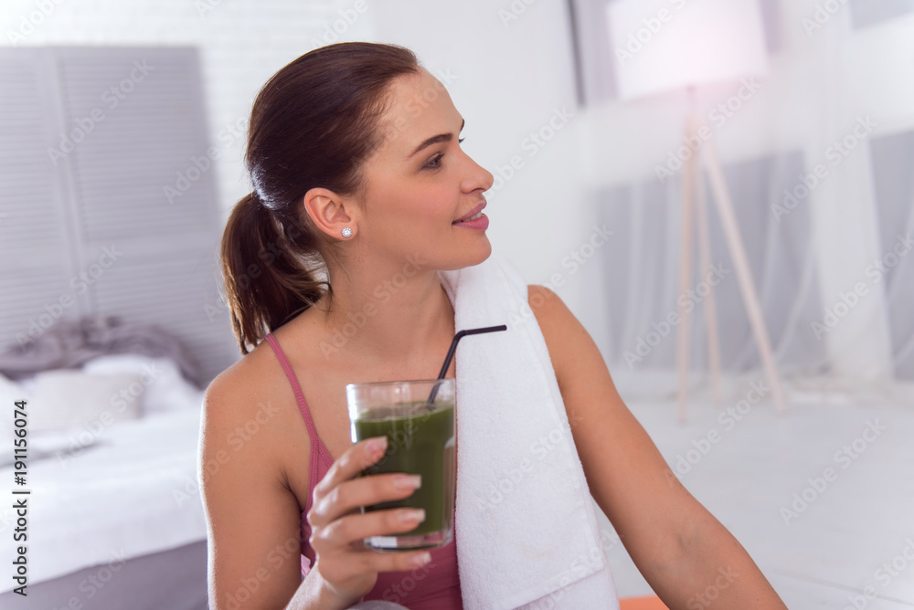 Relaxing. Beautiful smiling athletic dark-haired young woman holding a glass of fresh juice while sitting with a towel on her shoulder and looking in the distance Relaxing. Beautiful smiling athletic dark-haired young woman holding a glass of fresh juice while sitting with a towel on her shoulder and looking in the distance