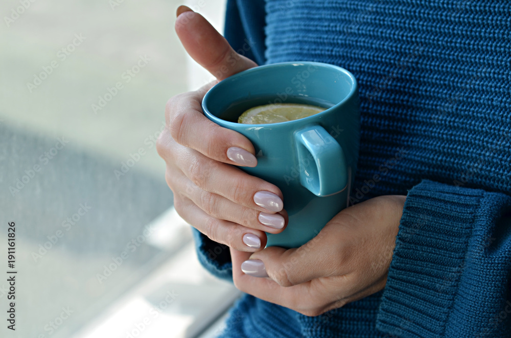 Woman in knitted sweater holding a cup of hot tea with lemon in hands with manicured nails. Turquoise and blue colors Woman in knitted sweater holding a cup of hot tea with lemon in hands with manicured nails. Turquoise and blue colors