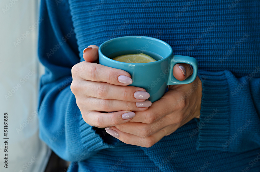 Woman in knitted sweater holding a cup of hot tea with lemon in hands with manicured nails. Turquoise and blue colors Woman in knitted sweater holding a cup of hot tea with lemon in hands with manicured nails. Turquoise and blue colors