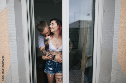 young man hugging and kissing beautiful smiling girlfriend in denim shorts and bra standing near window