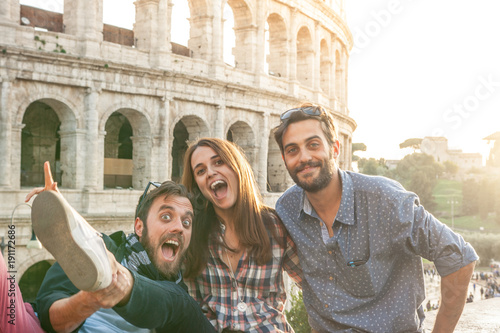 Three young friends tourists in front of colosseum in rome taking funny selfie pictures with smartphone camera. Sunset with lens flare.