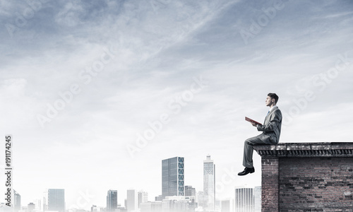 Wallpaper Mural Man on roof edge reading book and cityscape at background Torontodigital.ca