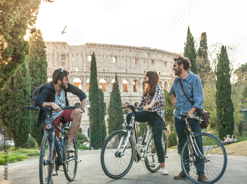 Three young friends tourists with bikes in colle oppio park in front of colosseum on road with trees at sunset having fun talking laughing in Rome lens flare