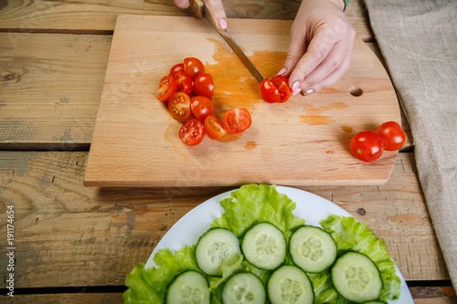 On a wooden table, a woman knifes a regimen of cherry tomatoes on slices for vegetable salad. Front view