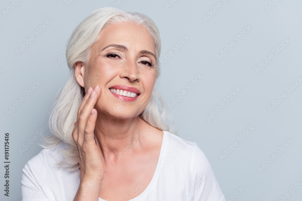 Charming, pretty, old woman touching her perfect soft face skin with fingers, smiling at camera over gray background, using day, night face cream, cosmetology procedures - obrazy, fototapety, plakaty 