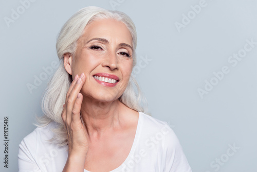 Charming, pretty, old woman touching her perfect soft face skin with fingers, smiling at camera over gray background, using day, night face cream, cosmetology procedures