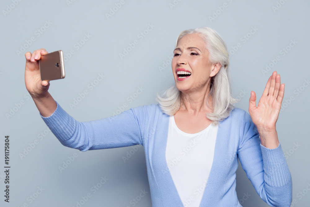 Obraz premium Portrait of excited happy cheerful delightful joyful charming mature lady granny tourist using her smatphone for video call, she is waving with a hand, isolated on gray background