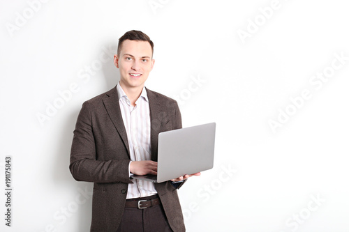 Smiling confident young man, no tie, holding grey laptop device and typing while standing against solid white wall. Wireless internet, wifi, enter password. Shaved, no facial hair. Isolated background