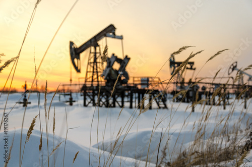 Blurred pump jack, wellhead and pipeline during sunset in the oilfield. Winter period. Oil and gas concept. Grass foreground. 