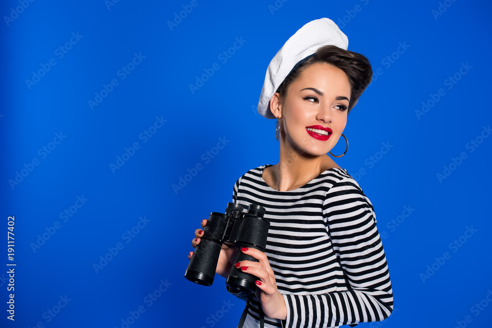 portrait of smiling young woman in retro clothing with binoculars isolated on blue