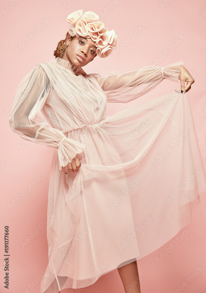 Soft fashion portrait of attractive black woman with light hair and green eyes wearing powder pink tulle dress and big frill hairband and posing. Pink background Soft fashion portrait of attractive black woman with light hair and green eyes wearing powder pink tulle dress and big frill hairband and posing. Pink background