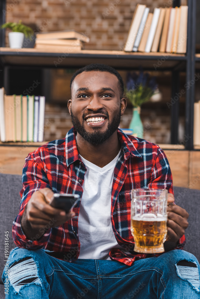 cheerful young african american man holding glass of beer and remote controller at home cheerful young african american man holding glass of beer and remote controller at home