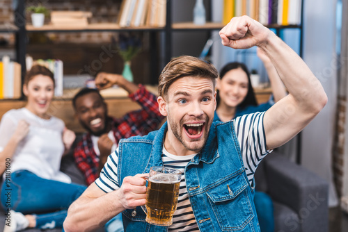 excited young man holding glass of beer and watching tv with friends sitting behind