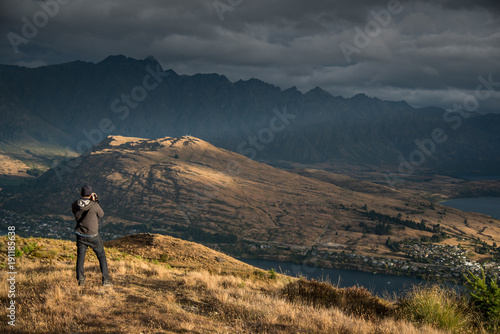 Young male photographer taking photo in photographing posture with mountain scenery during golden hour sunset in Queenstown, South Island, New Zealand. Travel and photography concept