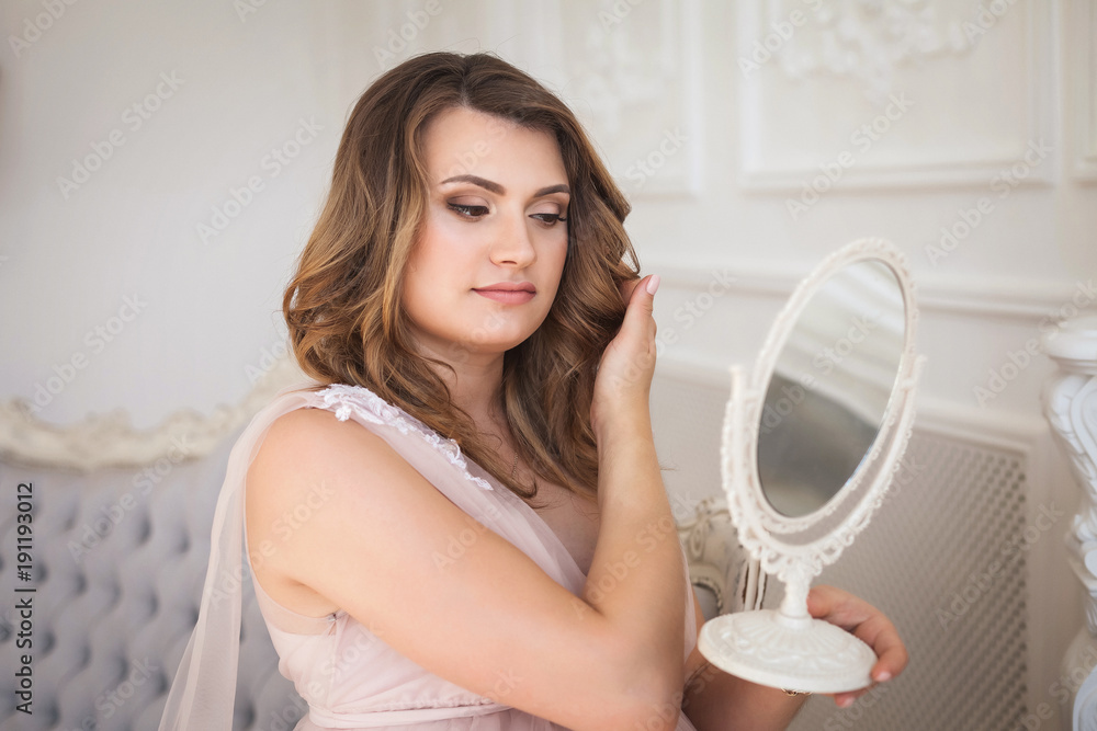 Young happy pregnant woman with curly hair in long elegant dress admiring the reflection in the mirror on vintage gray sofa in studio Young happy pregnant woman with curly hair in long elegant dress admiring the reflection in the mirror on vintage gray sofa in studio