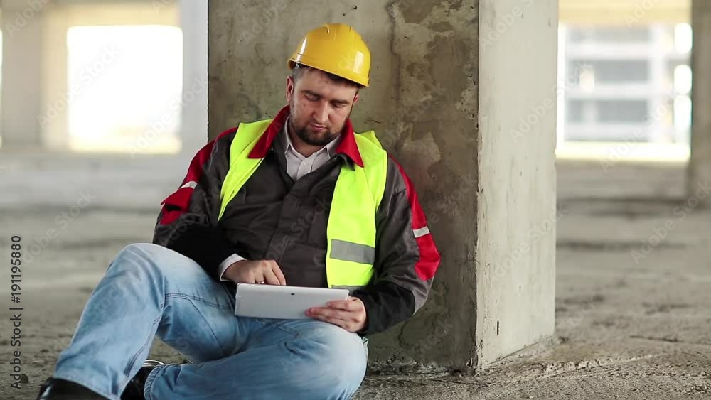 Workman with tablet computer sits on the floor. Worker work with tablet pc on construction site. Builder in white hard hat sits on concrete floor and holds in hands personal computer, looks in display Workman with tablet computer sits on the floor. Worker work with tablet pc on construction site. Builder in white hard hat sits on concrete floor and holds in hands personal computer, looks in display